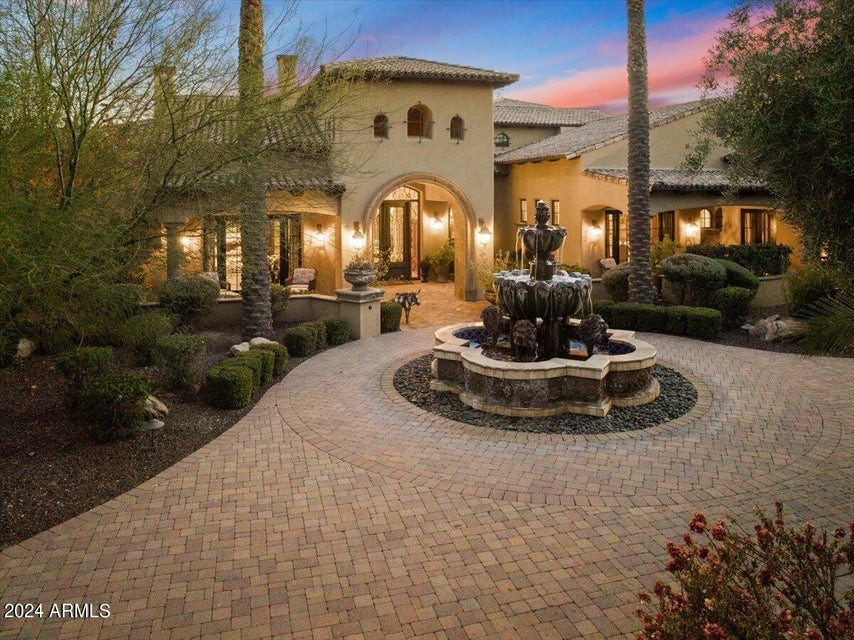 Front view at dusk of 9965 E Cholla Street in Scottsdale, a Mediterranean-style estate with a tiered stone fountain, circular drive and warm interior light under a twilight sky