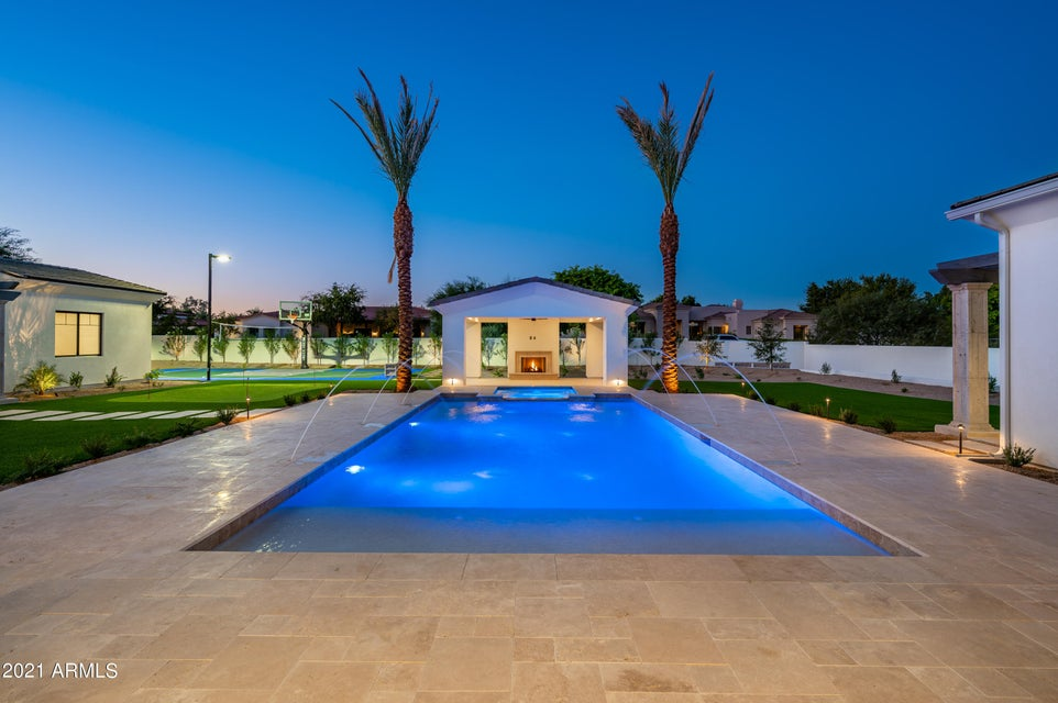Twilight backyard view of 6940 E Sunnyvale Rd in Paradise Valley, showing a glowing pool with arching jets, a white cabana with a lit fireplace and palm trees under a deep blue sky