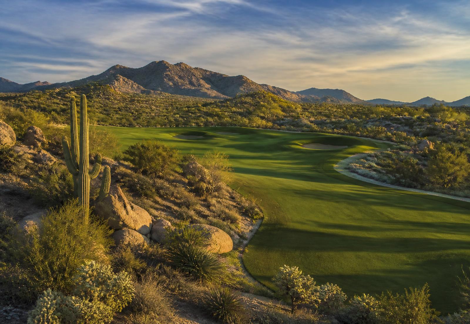 Championship desert golf fairway in the high Sonoran Desert of North Scottsdale in late afternoon light, edged by granite boulders, saguaro cactus and native desert with mountains beyond