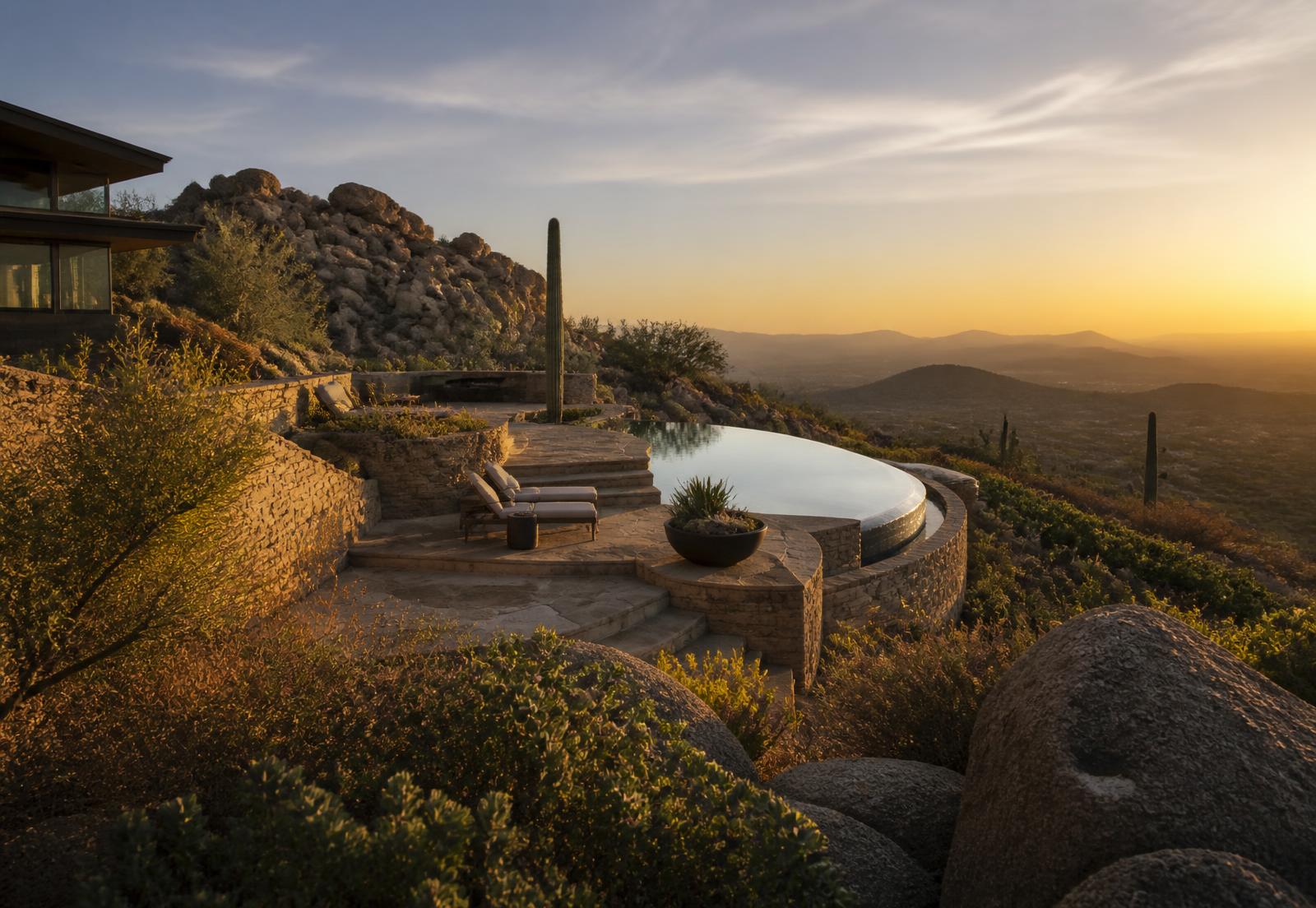 Elevated North Scottsdale estate terrace at twilight with an infinity-edge pool, granite boulders and saguaro overlooking layered desert ridgelines in warm golden light