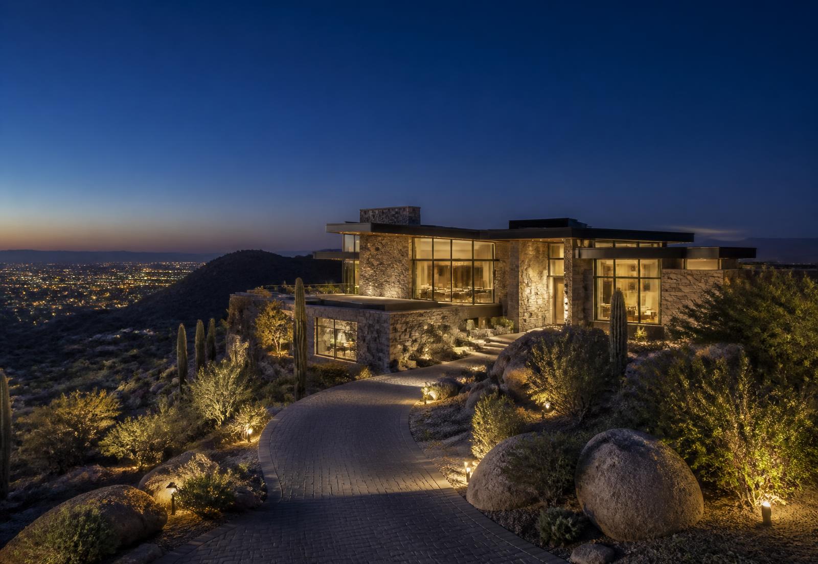 Contemporary North Scottsdale desert estate at twilight on an elevated homesite, warm interior light behind walls of glass, stone walls and a curved private driveway above distant valley city lights